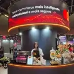Barista da Bom Café Eventos sorrindo atrás de um balcão de cafeteria circular escuro, iluminado por LED laranja, no luxuoso estande da Mastercard.