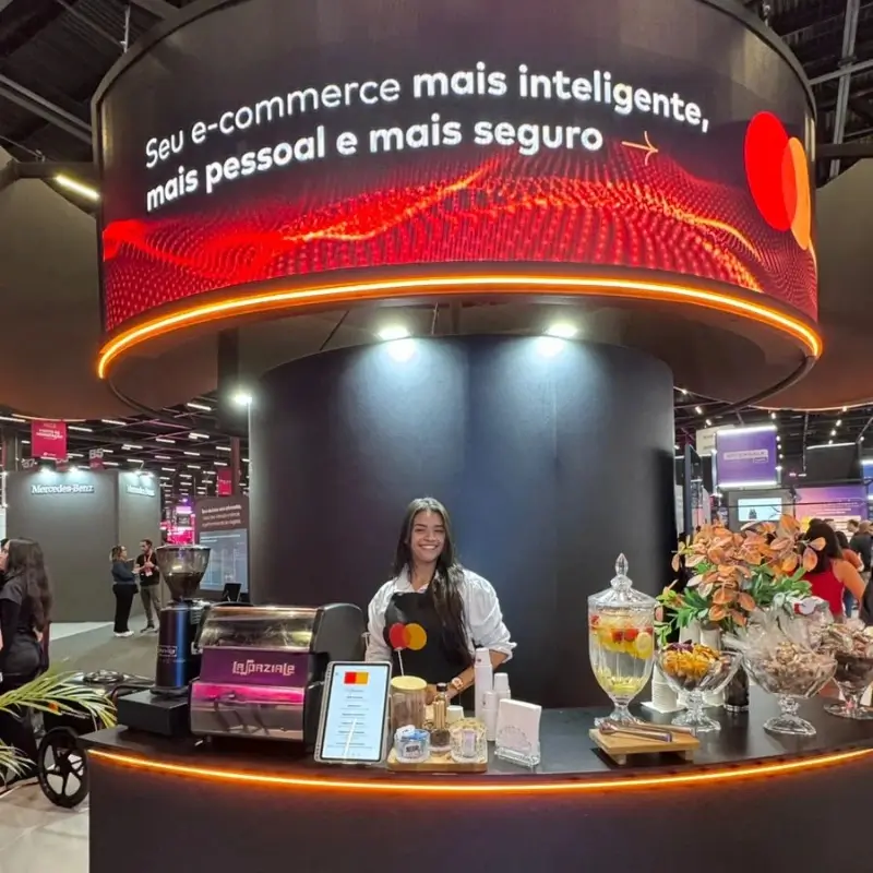 Barista da Bom Café Eventos sorrindo atrás de um balcão de cafeteria circular escuro, iluminado por LED laranja, no luxuoso estande da Mastercard.