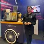 Barista da Bom Café Eventos com uniforme preto sorrindo em frente a um carrinho de café azul equipado com máquina de expresso no estande da marca Incentivare.