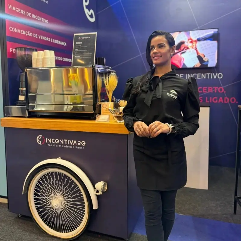 Barista da Bom Café Eventos com uniforme preto sorrindo em frente a um carrinho de café azul equipado com máquina de expresso no estande da marca Incentivare.