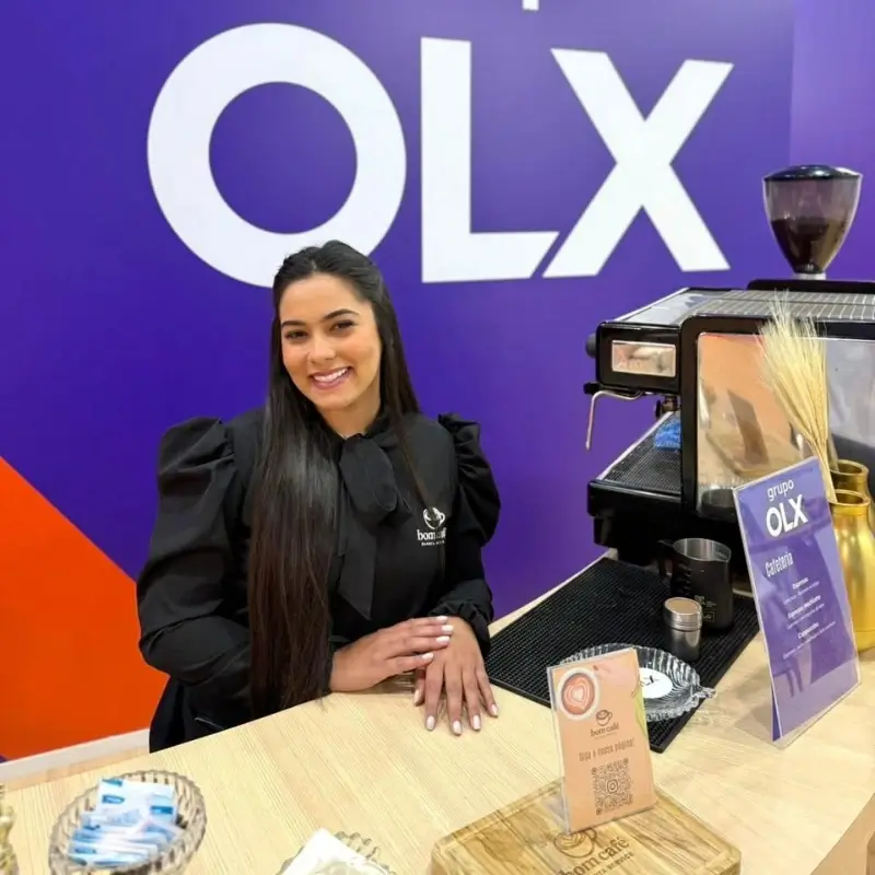 Barista da Bom Café Eventos com camisa preta sorrindo atrás do balcão de madeira no estande da OLX, com um grande painel roxo ao fundo