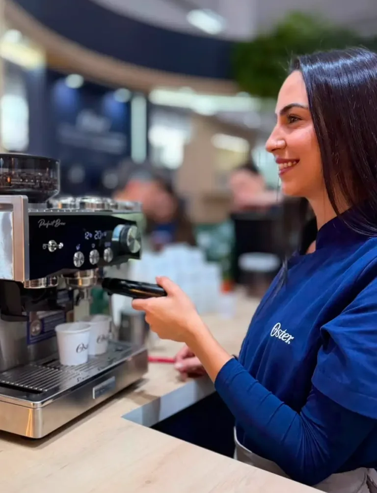 Barista de uniforme azul da Oster preparando café em máquina de espresso Perfect Brew em evento corporativo.