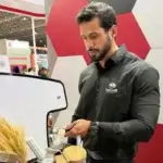 Barista homem da Bom Café Eventos concentrado, vestindo camisa preta com a logomarca da empresa, compactando café no porta-filtro da máquina