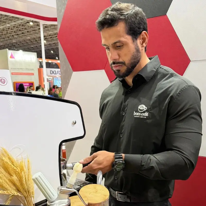 Barista homem da Bom Café Eventos concentrado, vestindo camisa preta com a logomarca da empresa, compactando café no porta-filtro da máquina