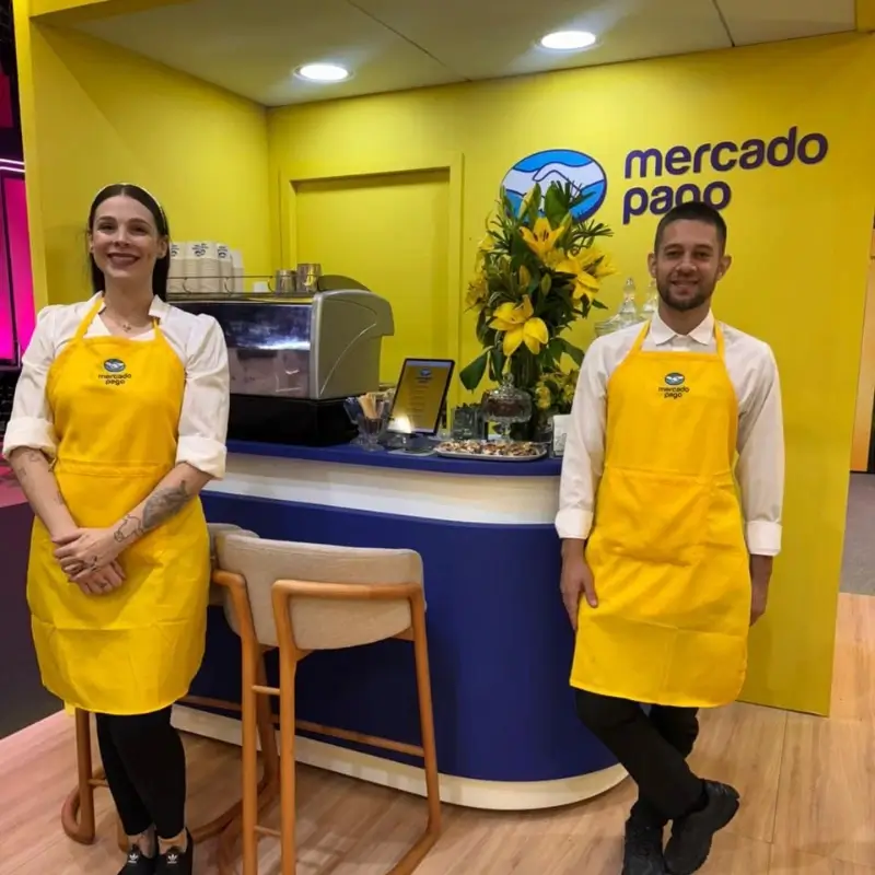 Dois baristas da Bom Café Eventos vestindo aventais amarelos com a logomarca do Mercado Pago, posicionados no balcão de atendimento do estande.