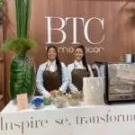Duas baristas da Bom Café Eventos de avental marrom sorrindo atrás do balcão de café branco e moderno no estande da BTC home decor.