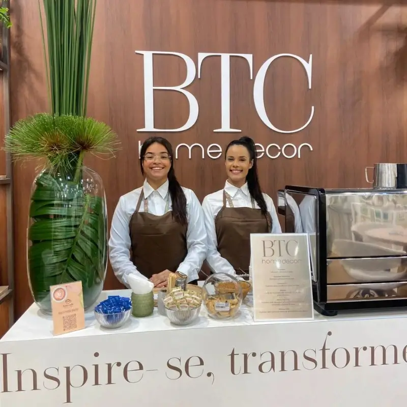 Duas baristas da Bom Café Eventos de avental marrom sorrindo atrás do balcão de café branco e moderno no estande da BTC home decor.