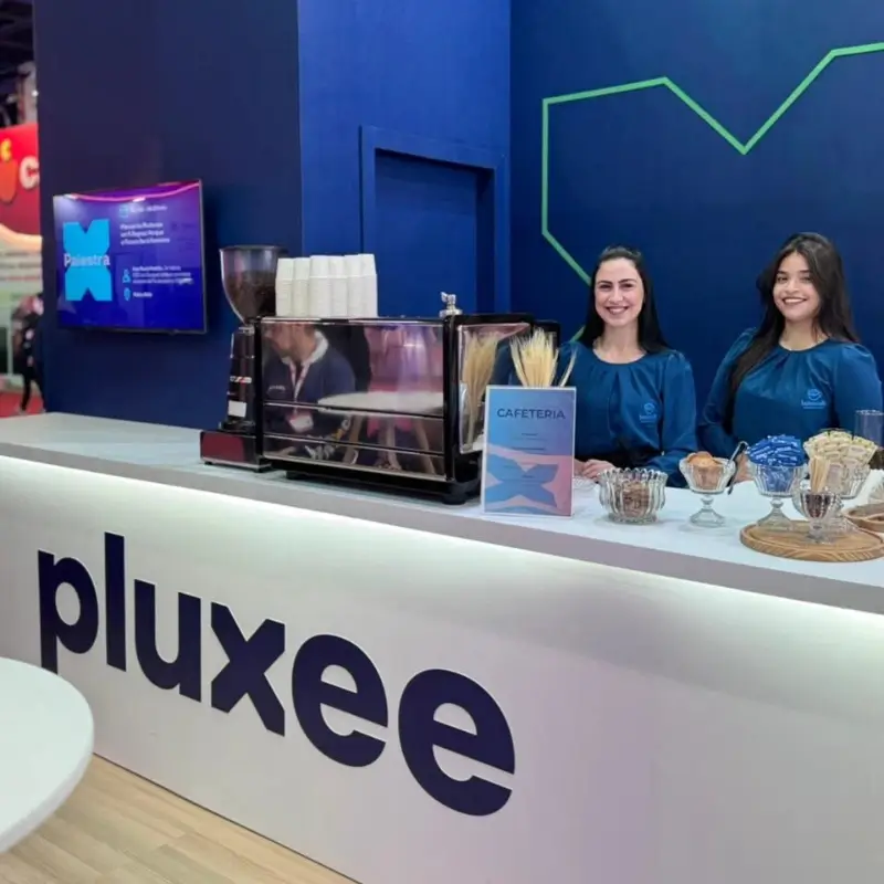 Duas baristas da Bom Café Eventos vestindo blusas azuis elegantes, sorrindo atrás do balcão de cafeteria branco no estande da Pluxee.