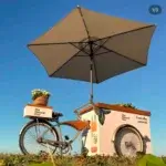Carrinho bike coffee ao ar livre com ombrelone bege e personalização da marca Natural One sob céu azul.