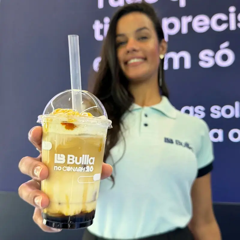 Copo transparente de café gelado com caramelo e logomarca "Bullla no CONARH" em destaque, segurado por uma barista da Bom Café Eventos desfocada ao fundo.
