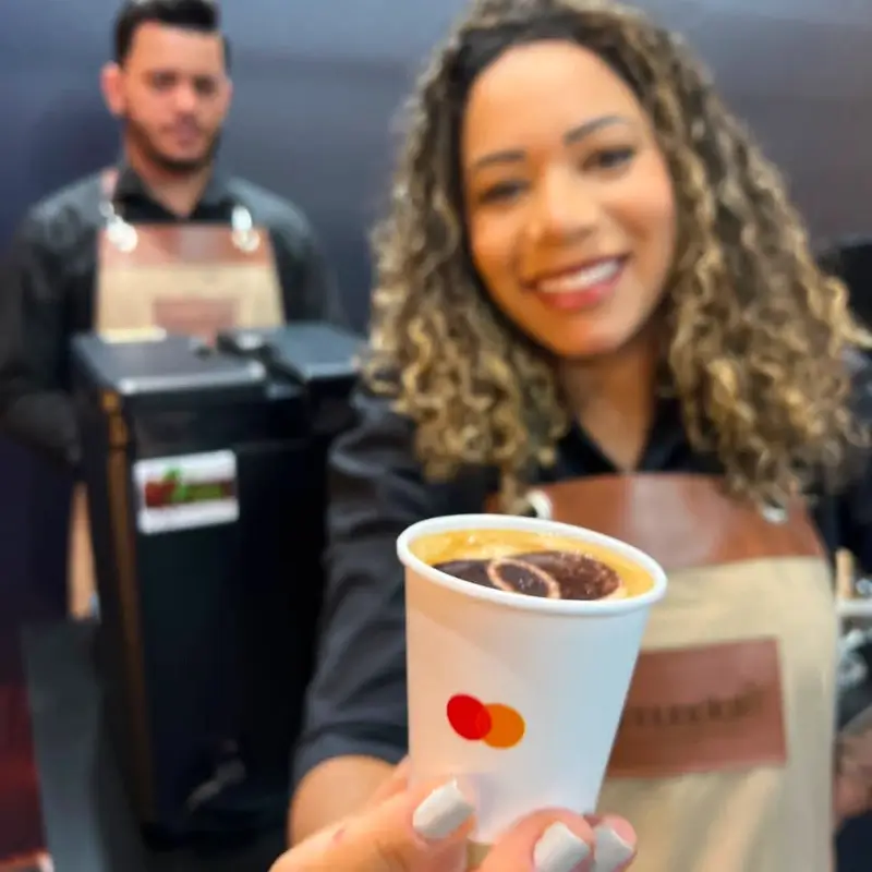 Barista da Bom Café Eventos em primeiro plano, levemente desfocada e sorrindo, oferecendo um copinho de papel com a logomarca da Mastercard, destacando a bebida no interior com arte em pó de cacau.