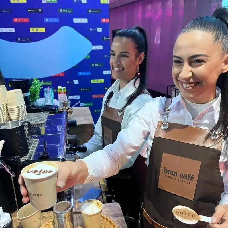 Duas baristas de avental de couro da Bom Café Eventos sorrindo e entregando um copo de cappuccino com a logomarca da Veloe impressa em pó de cacau.