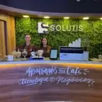 Duas baristas uniformizadas da Bom Café Eventos no balcão de madeira do estande da Solutis, com o letreiro "Apaixonados por Café, Tecnologia e Negócios".