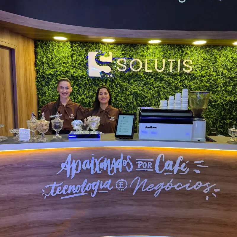 Duas baristas uniformizadas da Bom Café Eventos no balcão de madeira do estande da Solutis, com o letreiro "Apaixonados por Café, Tecnologia e Negócios".