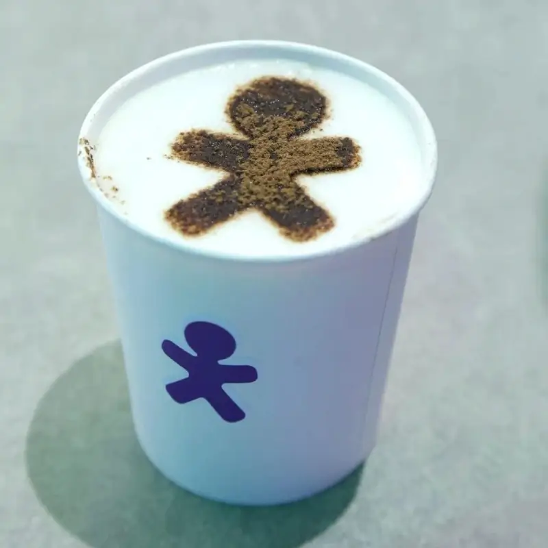 Copo de papel com a logomarca da Vivo, exibindo um cappuccino com a imagem do mascote da marca impressa em cacau sobre a espuma de leite, preparado pela Bom Café Eventos.