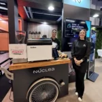 Baristas ao lado da estação bike de café personalizada Núclea em estande de feira corporativa.