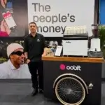 Barista de uniforme preto ao lado do carrinho bike de café personalizado Oobit em estande corporativo.