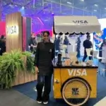 Barista da Bom Café Eventos de uniforme preto sorrindo ao lado de um carrinho de café em madeira rústica, personalizado com a logomarca da Visa em evento corporativo.