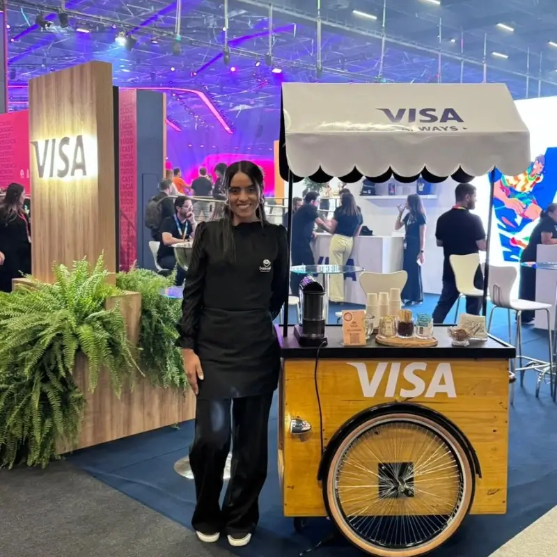 Barista da Bom Café Eventos de uniforme preto sorrindo ao lado de um carrinho de café em madeira rústica, personalizado com a logomarca da Visa em evento corporativo.
