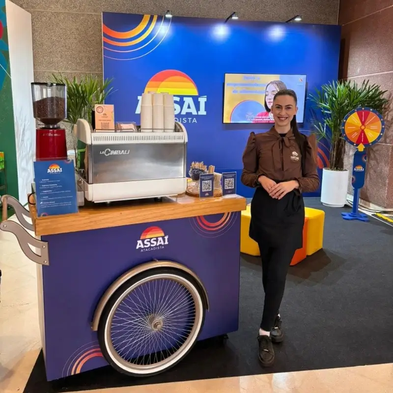 Barista da Bom Café Eventos com camisa marrom e avental preto sorrindo ao lado de um carrinho de café azul escuro adesivado com a logomarca do Assaí Atacadista, operando uma máquina de expresso La Cimbali branca.