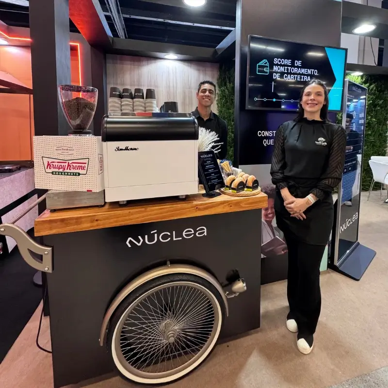 Dupla de baristas uniformizados da Bom Café Eventos sorrindo atrás de um carrinho de café estilo triciclo vintage. O balcão possui uma máquina de expresso profissional branca e uma caixa de donuts, montado no estande corporativo da marca Núclea.