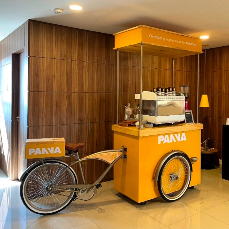 Carrinho de café tipo triciclo vintage (coffee bike) amarelo com detalhes em madeira, personalizado com a logomarca da PANVA. O carrinho possui um letreiro superior com a frase "Conexões que geram grandes histórias!" e uma máquina de expresso branca, estrutura montada pela Bom Café Eventos em um ambiente corporativo.