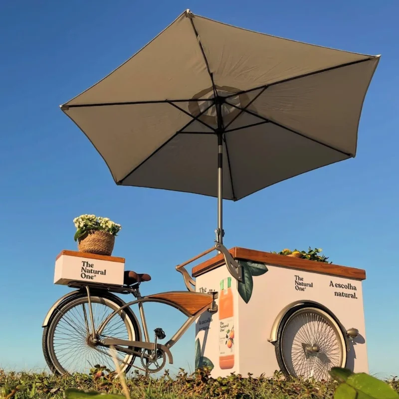 Coffee bike branca da Bom Café Eventos estacionada sobre o gramado ao ar livre sob um céu azul claro. O triciclo possui um grande guarda-sol bege, detalhes em madeira, um cesto de flores na garupa e é adesivado com a identidade visual da marca The Natural One.