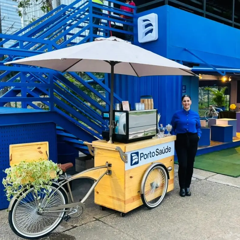 Barista da Bom Café Eventos vestindo camisa azul elegante, sorrindo ao lado de um carrinho de café estilo bicicleta em madeira clara com guarda-sol e logomarca da Porto Saúde, posicionado em frente a contêineres azuis.
