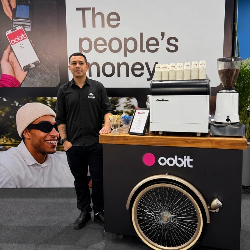 Barista profissional da Bom Café Eventos de camisa preta posando ao lado de um carrinho de café estilo triciclo (coffee bike) preto. O balcão é adesivado com a logomarca da Oobit e equipado com uma máquina de expresso branca da San Marco e moinho de grãos, montado em um estande corporativo.