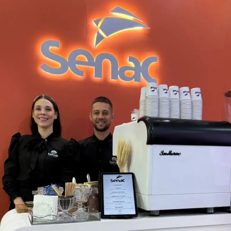 Dupla de baristas da Bom Café Eventos com uniformes pretos padronizados sorrindo no balcão de café expresso, com a logomarca iluminada do Senac ao fundo.
