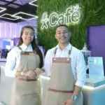 Dupla de baristas profissionais da Bom Café Eventos sorrindo atrás de um balcão moderno de cafeteria. Eles vestem camisas brancas e aventais beges elegantes com alças de couro. Ao fundo, um painel de folhagens com um letreiro luminoso em neon no estande da Vivo.