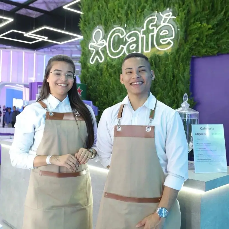 Dupla de baristas profissionais da Bom Café Eventos sorrindo atrás de um balcão moderno de cafeteria. Eles vestem camisas brancas e aventais beges elegantes com alças de couro. Ao fundo, um painel de folhagens com um letreiro luminoso em neon no estande da Vivo.