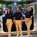 Três baristas da Bom Café Eventos usando uniformes azuis da Oster alinhados em frente a um balcão de cafeteria circular iluminado.