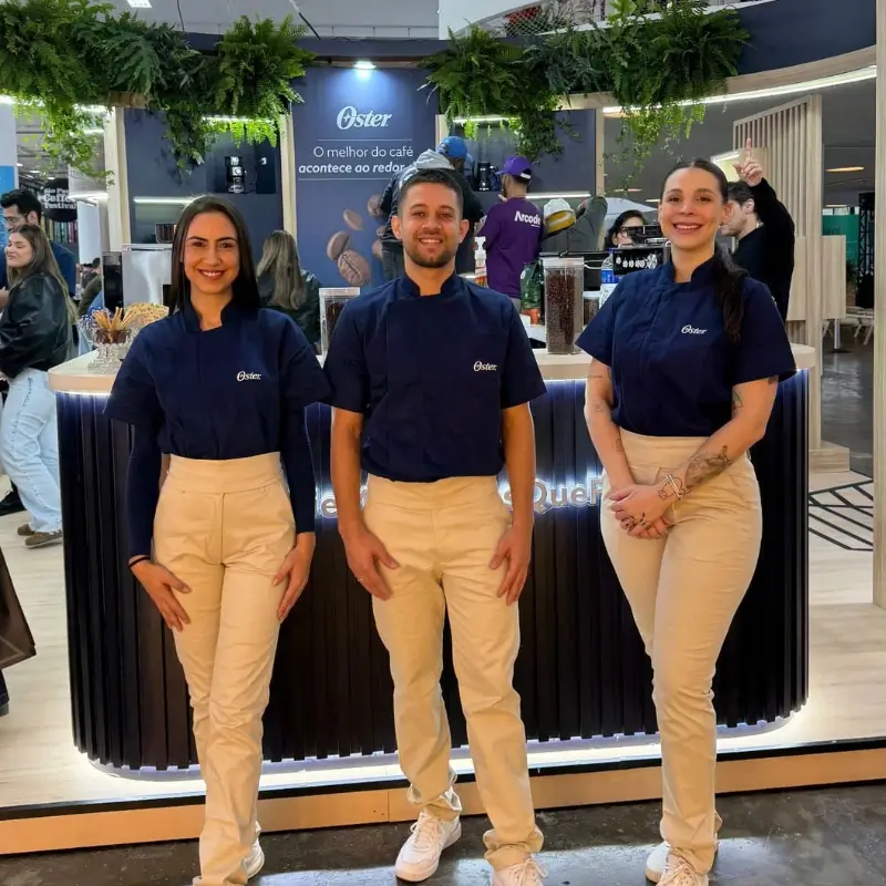 Três baristas da Bom Café Eventos usando uniformes azuis da Oster alinhados em frente a um balcão de cafeteria circular iluminado.