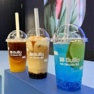 Três copos transparentes alinhados em um balcão azul com bebidas refrescantes: um chá gelado, um iced coffee cremoso com caramelo e uma soda italiana azul com limão. Todos os copos possuem a logomarca da "Bullla no CONARH 26", preparados pela Bom Café Eventos.