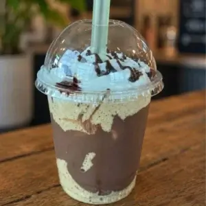 Copo transparente sobre uma mesa de madeira contendo um frapê de café cremoso, decorado com calda de chocolate nas laterais e uma generosa cobertura de chantilly, uma das especialidades da Bom Café Eventos.