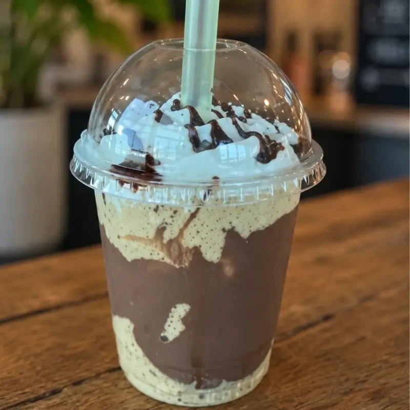 Copo transparente sobre uma mesa de madeira contendo um frapê de café cremoso, decorado com calda de chocolate nas laterais e uma generosa cobertura de chantilly, uma das especialidades da Bom Café Eventos.