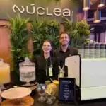 Dupla de baristas da Bom Café Eventos sorrindo no balcão do estande da Núclea ao lado de uma máquina de café expresso Sanremo.