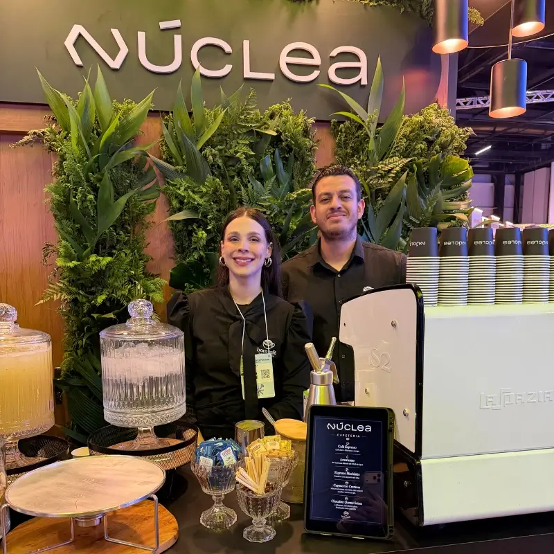 Dupla de baristas da Bom Café Eventos sorrindo no balcão do estande da Núclea ao lado de uma máquina de café expresso Sanremo.