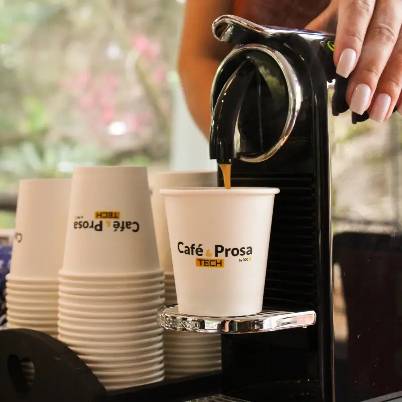 Mão de uma mulher operando uma máquina de café em cápsulas, extraindo um café expresso diretamente em um copo de papel branco personalizado com a marca do cliente, em serviço organizado pela Bom Café Eventos.