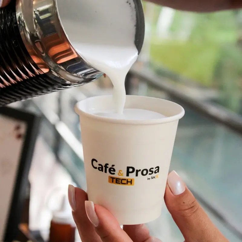 Mão segurando um copo de papel sustentável personalizado do Café e Prosa Tech recebendo creme de leite vaporizado de uma jarra manuseada pela equipe da Bom Café Eventos.